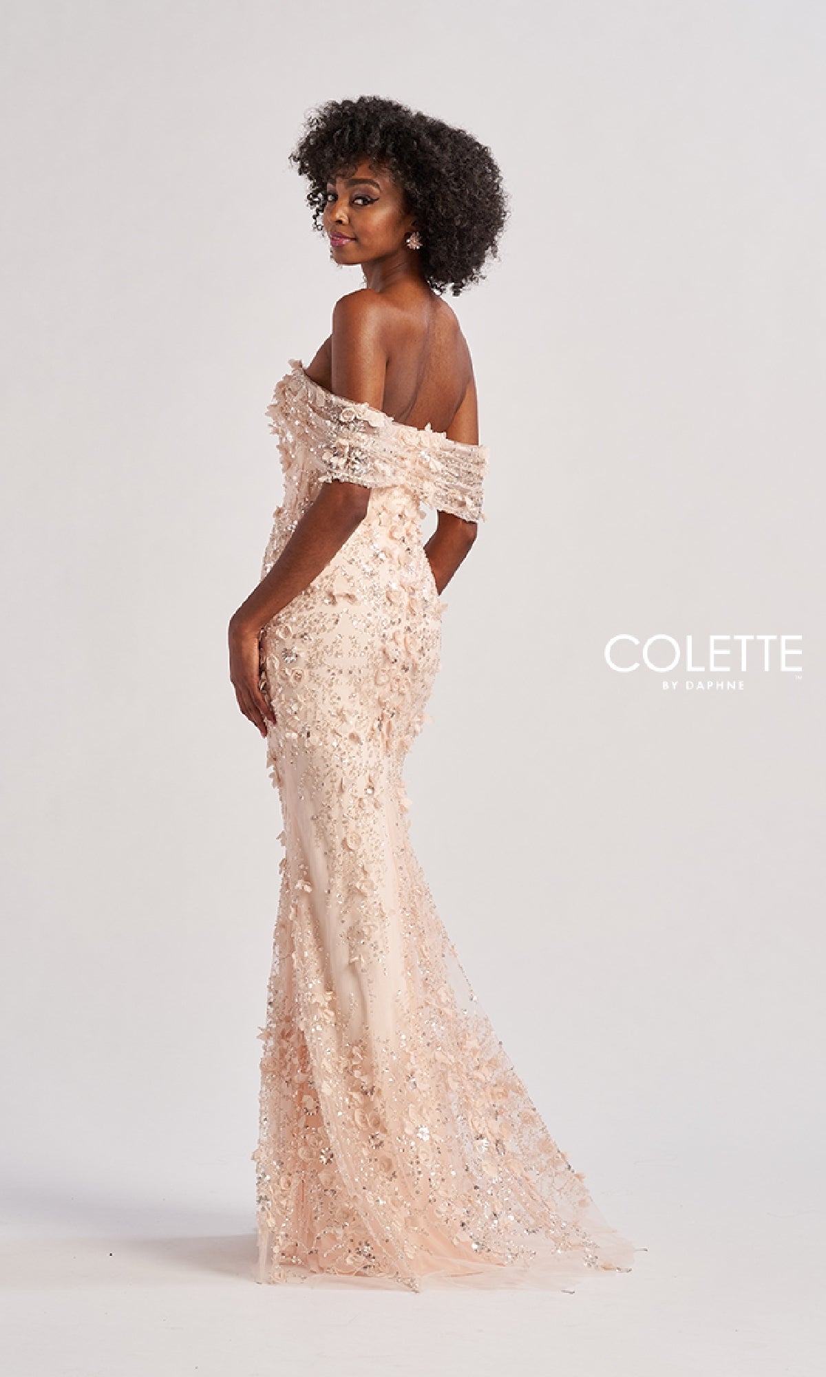 A woman poses against a white background wearing the Colette CL8530, a blush pink, off-the-shoulder long prom dress with sequins, floral embellishments, and a fitted silhouette.
